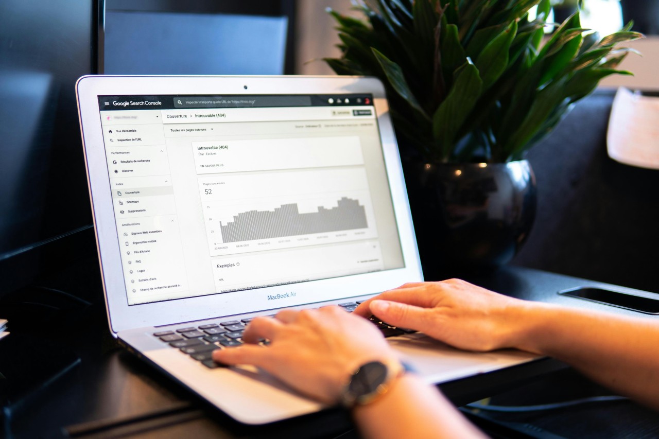 Person's hands typing on a MacBook Air displaying Google Search Console analytics dashboard. The screen shows SEO performance data including graphs, metrics, and navigation menu on the left sidebar. A green plant sits in the background on a modern desk setup, creating a professional work environment.